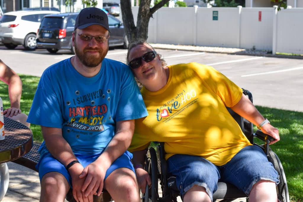 smiling married couple seated outdoors, one at bench, one in wheelchair