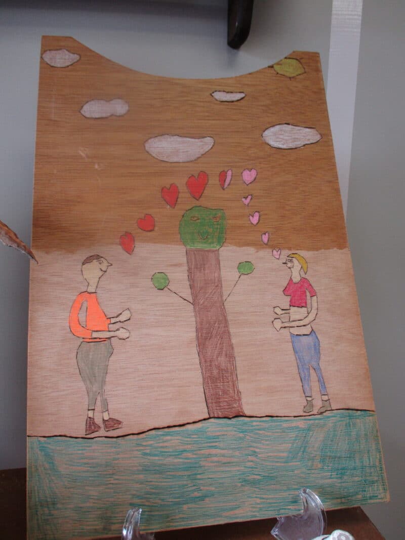 artwork wood carving of man and woman looking at each other with a tree in between them, with hearts and clouds above them