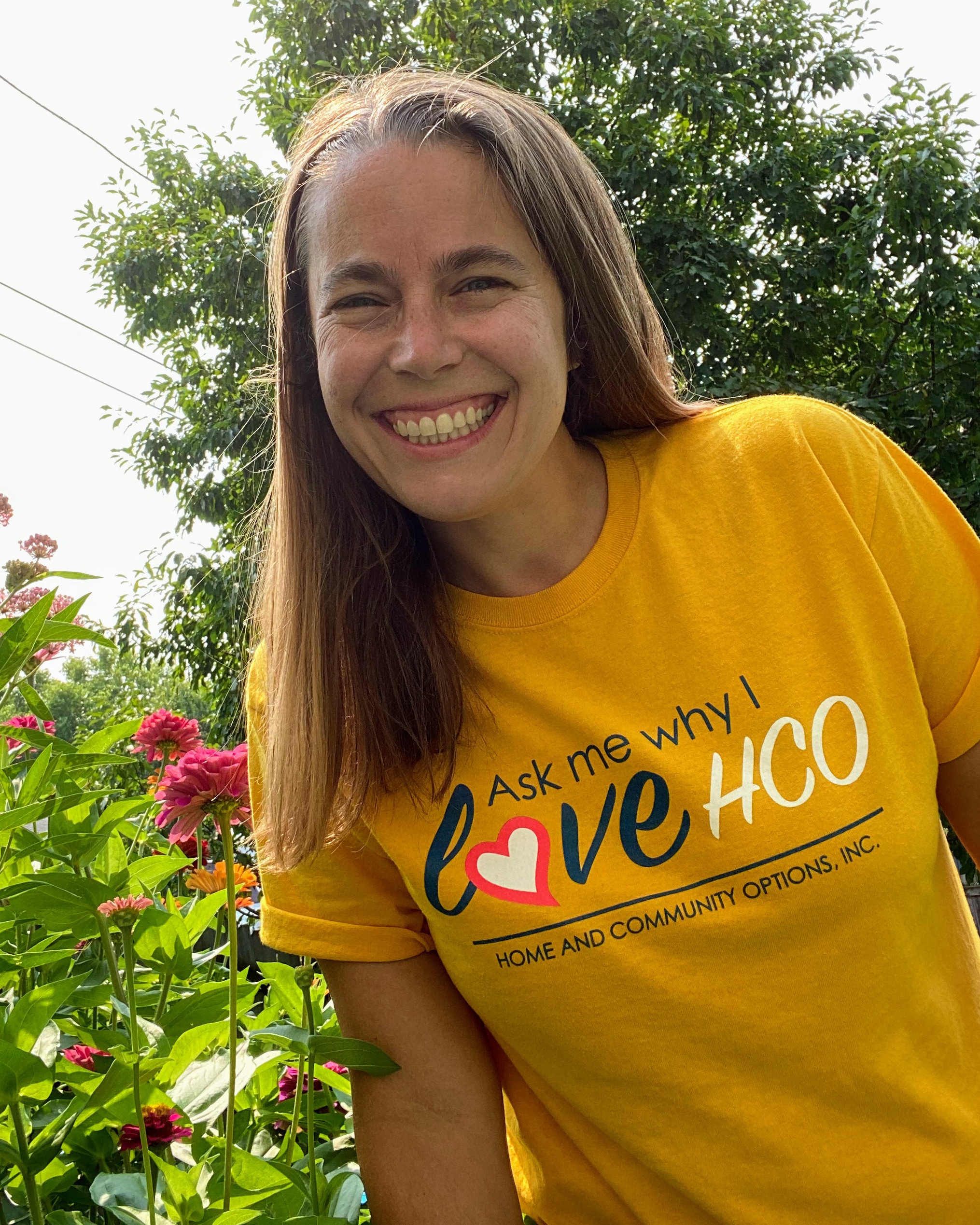 Smiling woman standing next to flowers wearing t-shirt saying ask me why I love HCO: Home and Community Options
