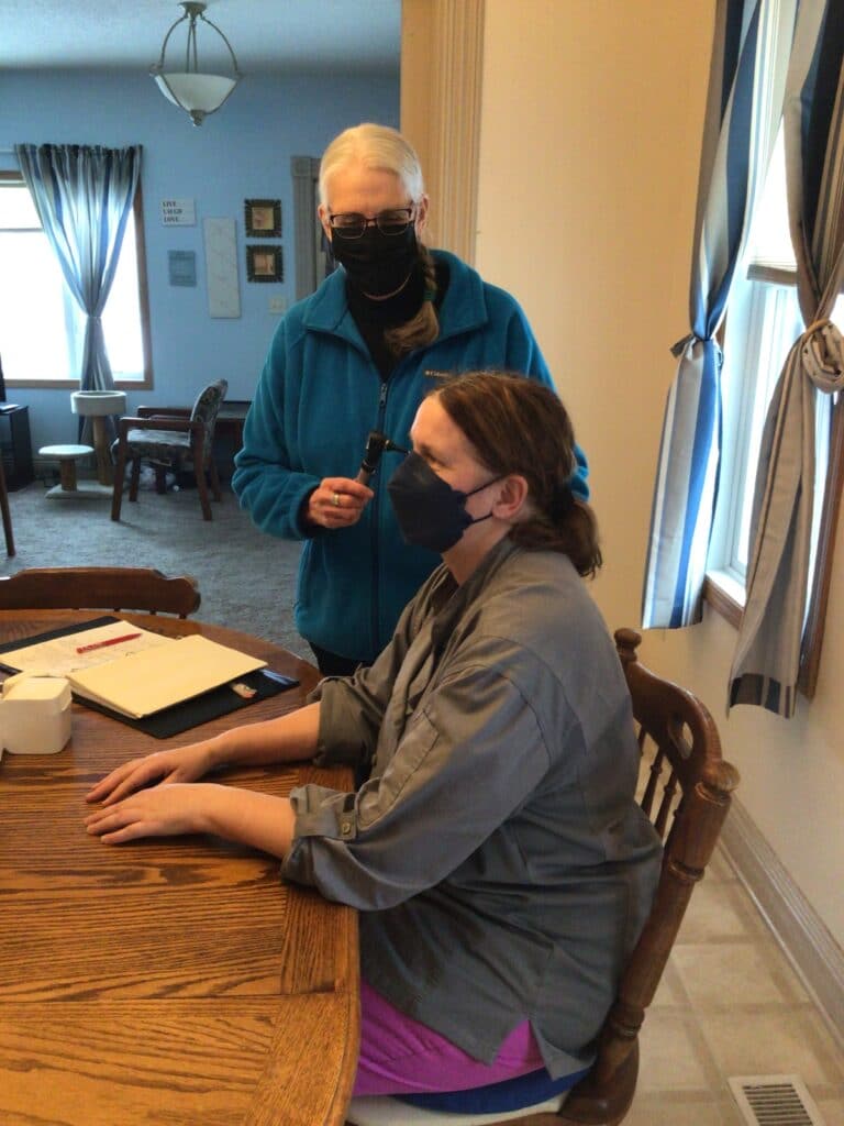 Two masked women, one seated at dining room table, the other is a nurse holding an otoscope to look inside the seated woman's ear