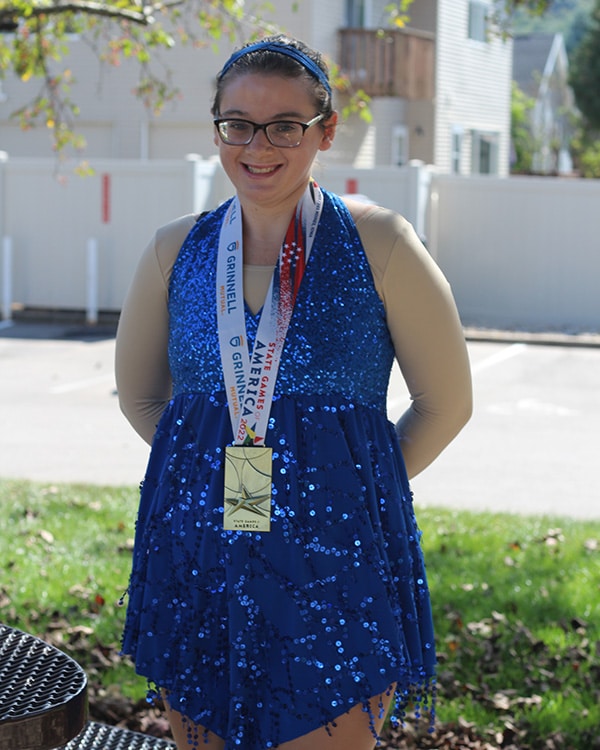 Smiling woman in blue sequins ice skating costume wearing gold medal skating award with a star and text "state games of America"